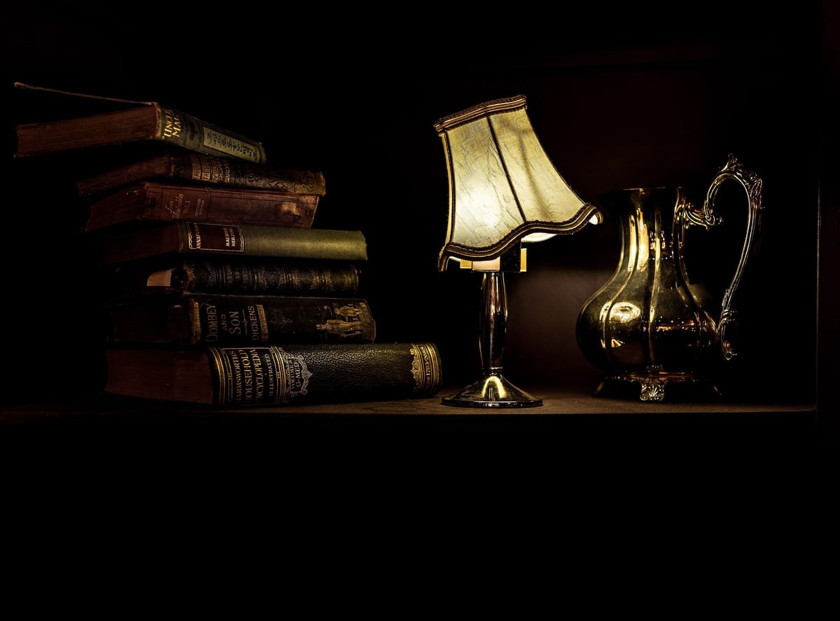 De vieux livres reliés empilés dans la pénombre, à côté d’une petite lampe allumée et d’un pichet en métal.