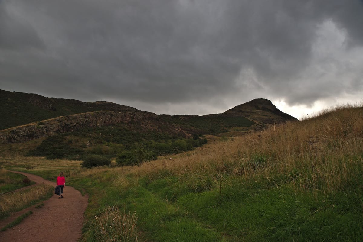 Sous un ciel d’orage gris ardoise, une silhouette en veste rouge marche seule sur un sentier de terre qui serpente entre les herbes hautes, vers une colline sombre.