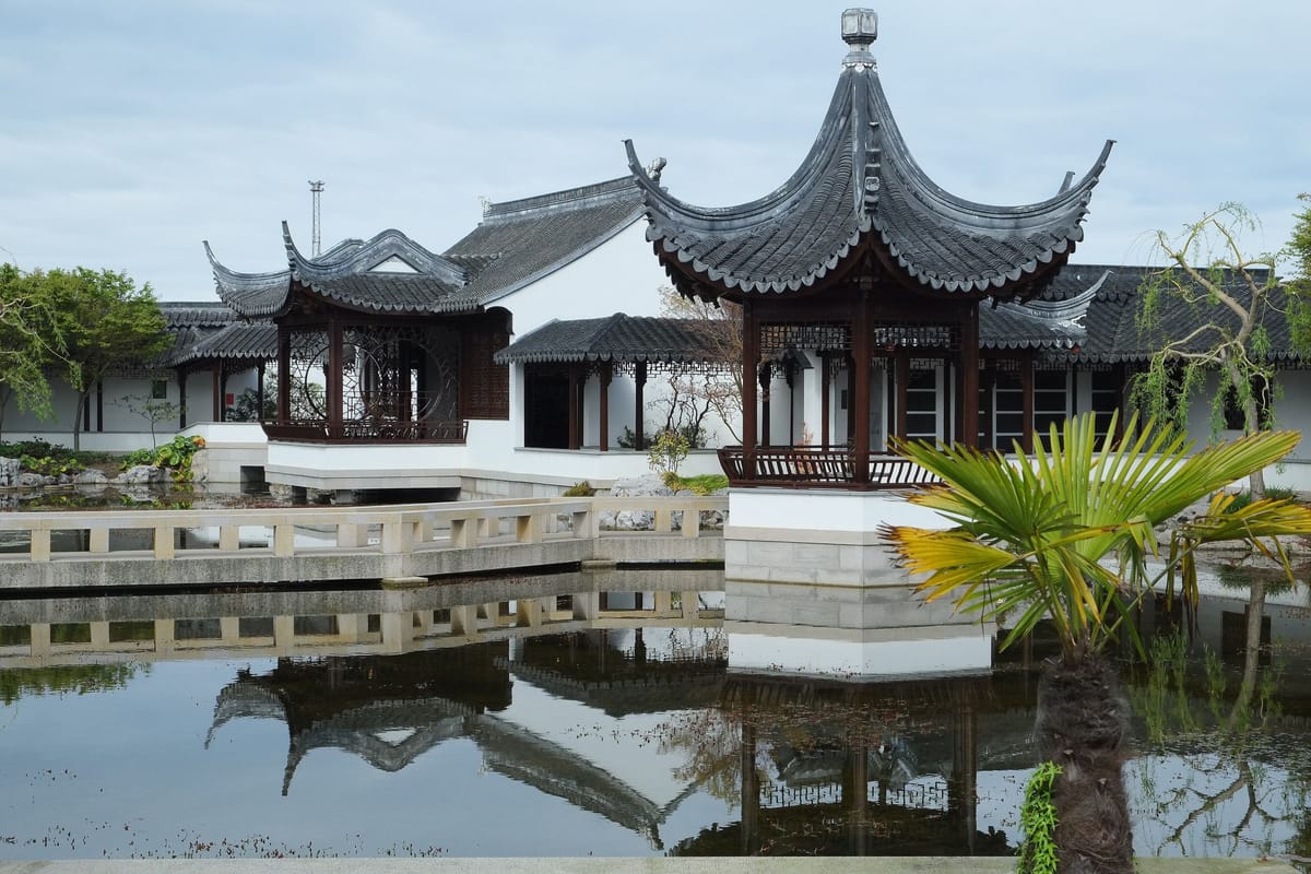 Un pavillon chinois aux toits recourbés se reflète dans l’eau calme d’un bassin, devant des murs blancs et des arbres, dans un jardin de style classique.