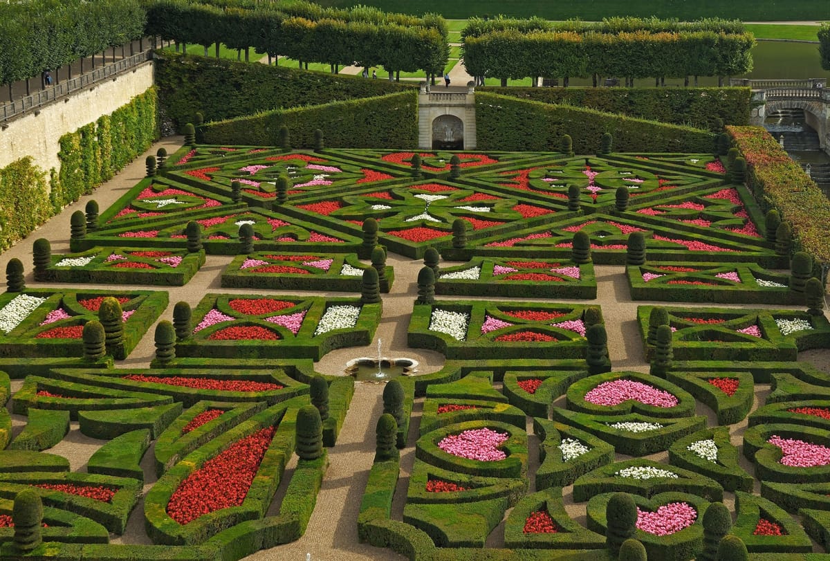 Vu d’en haut, un jardin à la française aux parterres de buis taillés en motifs symétriques, garnis de fleurs rouges, roses et blanches, autour d’une petite fontaine.