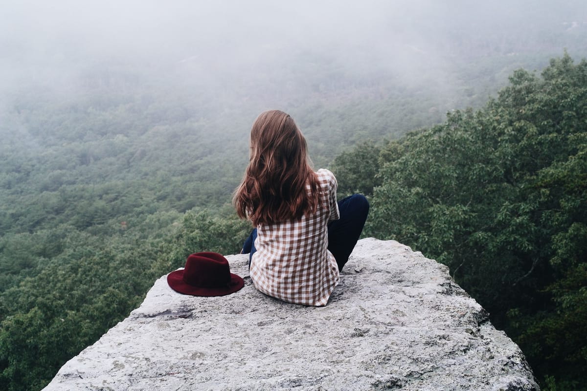 Vue de dos, une jeune femme aux longs cheveux châtains est assise en tailleur au bord d’un rocher, face à une vallée boisée noyée dans la brume ; un chapeau bordeaux est posé à côté d’elle.