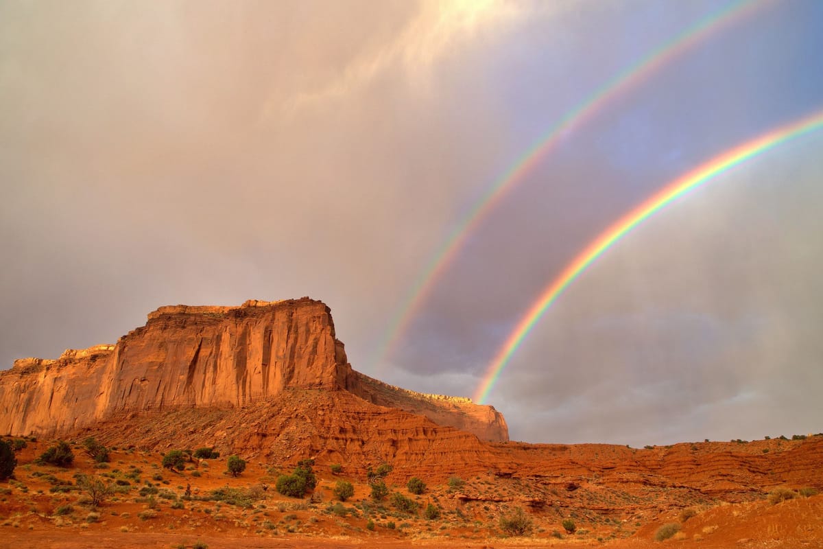 Un double arc-en-ciel se déploie dans un ciel d’orage rosé au-dessus d’une haute falaise de grès rouge et d’une plaine désertique semée de buissons.