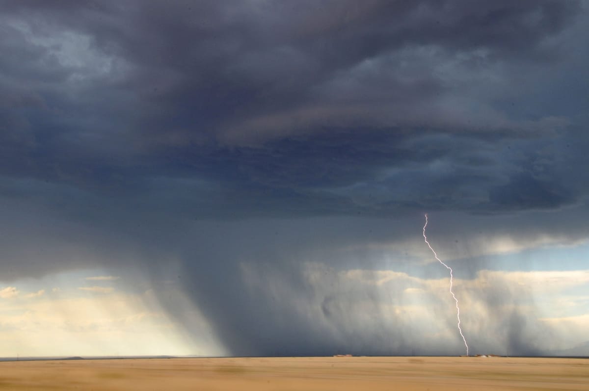Sous un ciel d’orage bleu ardoise, un rideau de pluie tombe sur une vaste plaine blonde et un éclair frappe le sol à l’horizon.